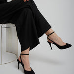 Person wearing black high-heeled shoes and pants sitting on a chair against a plain background