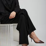 Person wearing a black outfit with high-heeled shoes sitting on a wire stool against a white background