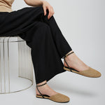 Person wearing beige flats and black pants sitting on a wire stool against a white background