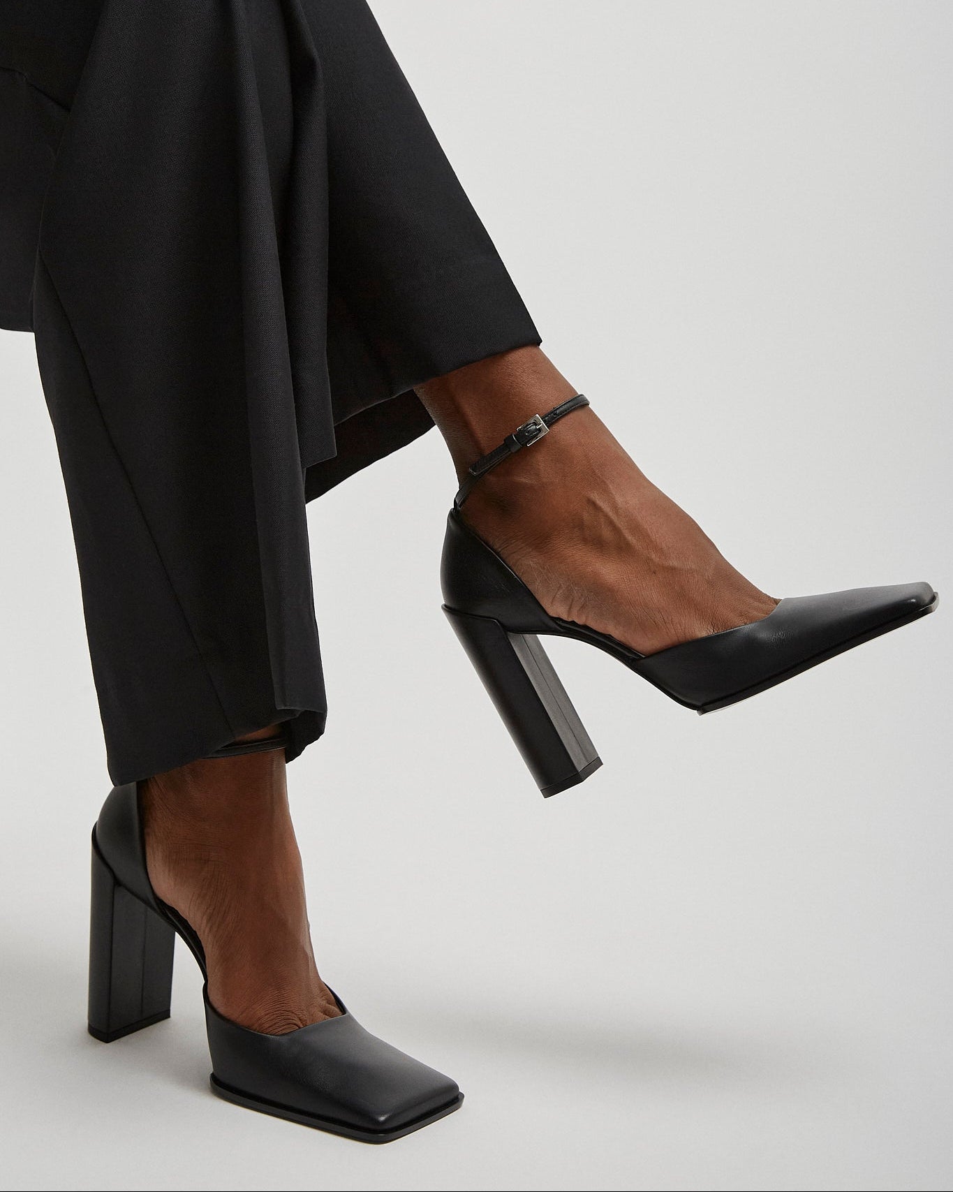 Black high-heeled shoes worn with black pants on a light gray background