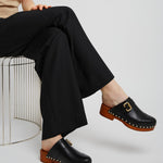 Person wearing black clogs with brown soles and a gold buckle, sitting on a wire chair.