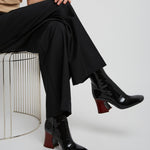 Person wearing black pants and black shoes sitting on a wire stool against a white background