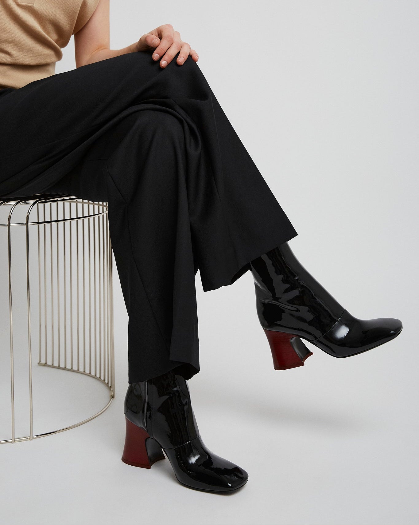 Person wearing black pants and black shoes sitting on a wire stool against a white background