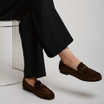 Person wearing dark brown loafers and black pants sitting on a white chair.