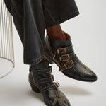 Black studded boots with gold buckles worn by a person sitting on a white chair.