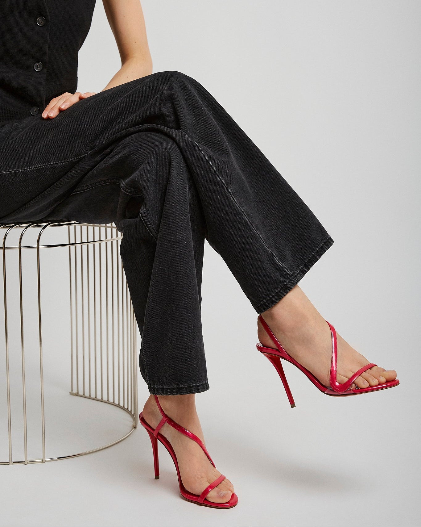 Person wearing black outfit with red high-heeled shoes sitting on a wire stool against a white background