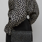 Person wearing a black and white polka dot blouse with a black skirt and black clutch bag on a gray background