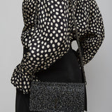Person wearing a black and white polka dot blouse with a black skirt and black clutch bag on a gray background