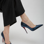 Navy high-heeled shoes with red soles worn by a person in black pants on a light gray background
