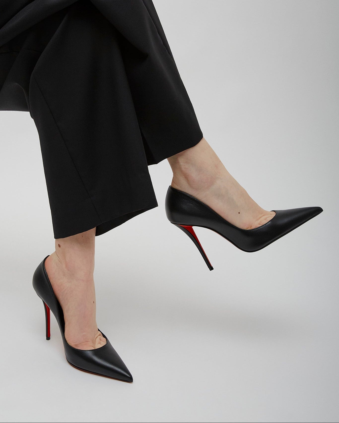 Person wearing black high-heeled shoes with red soles and black pants on a light gray background