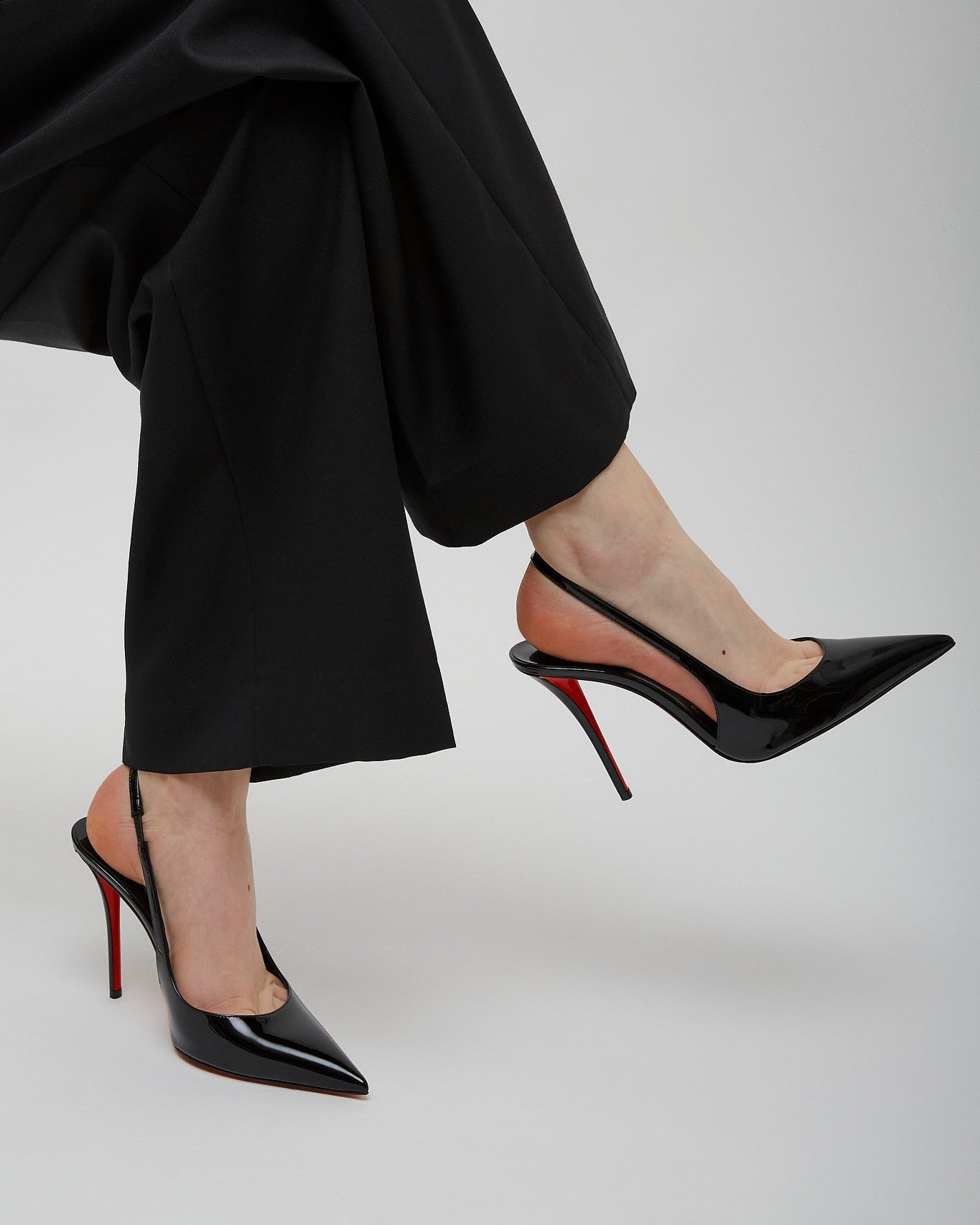 Black high-heeled shoes with red soles worn by a person in black pants on a light gray background