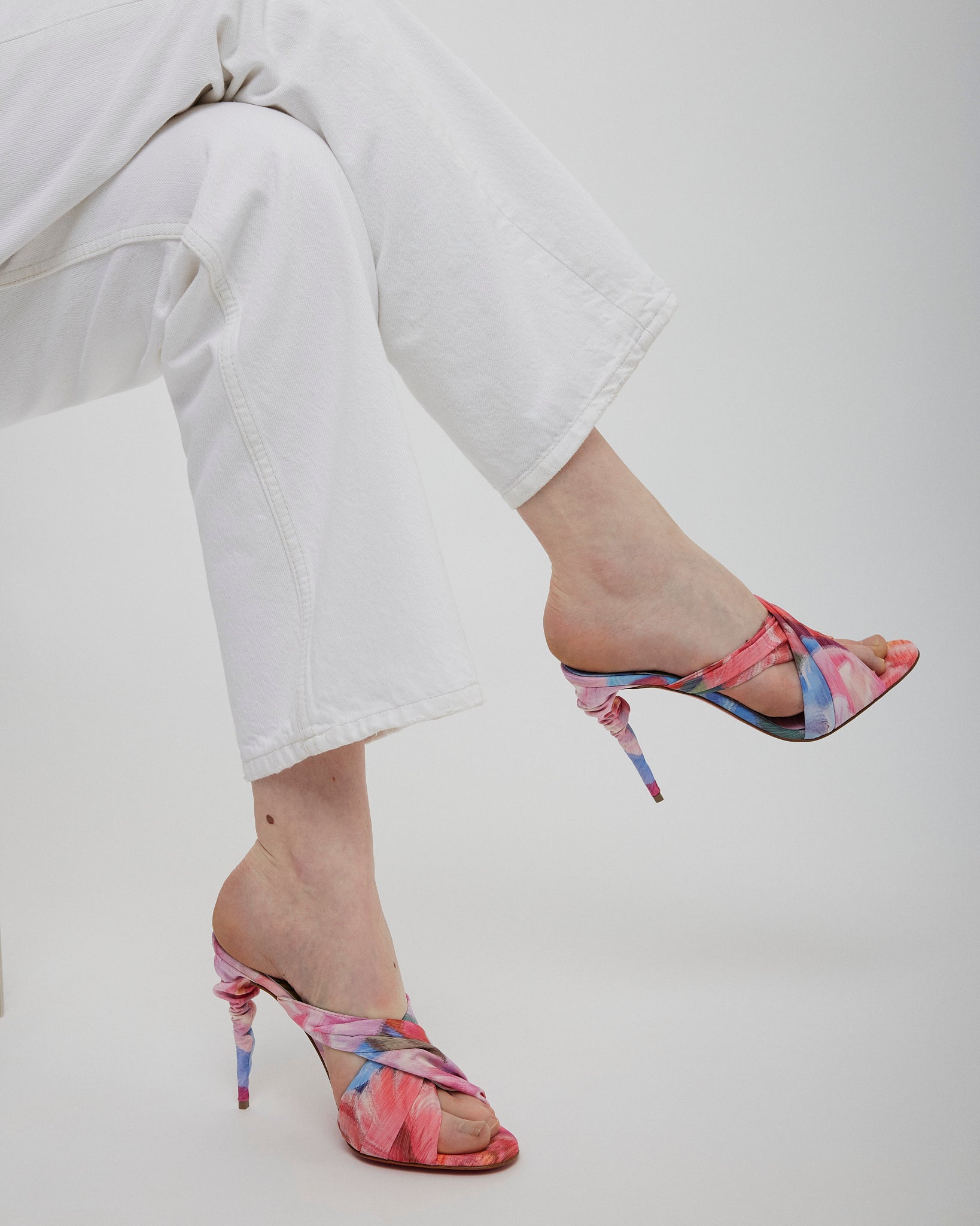 Multicolored high-heeled sandals worn with white pants on a light gray background