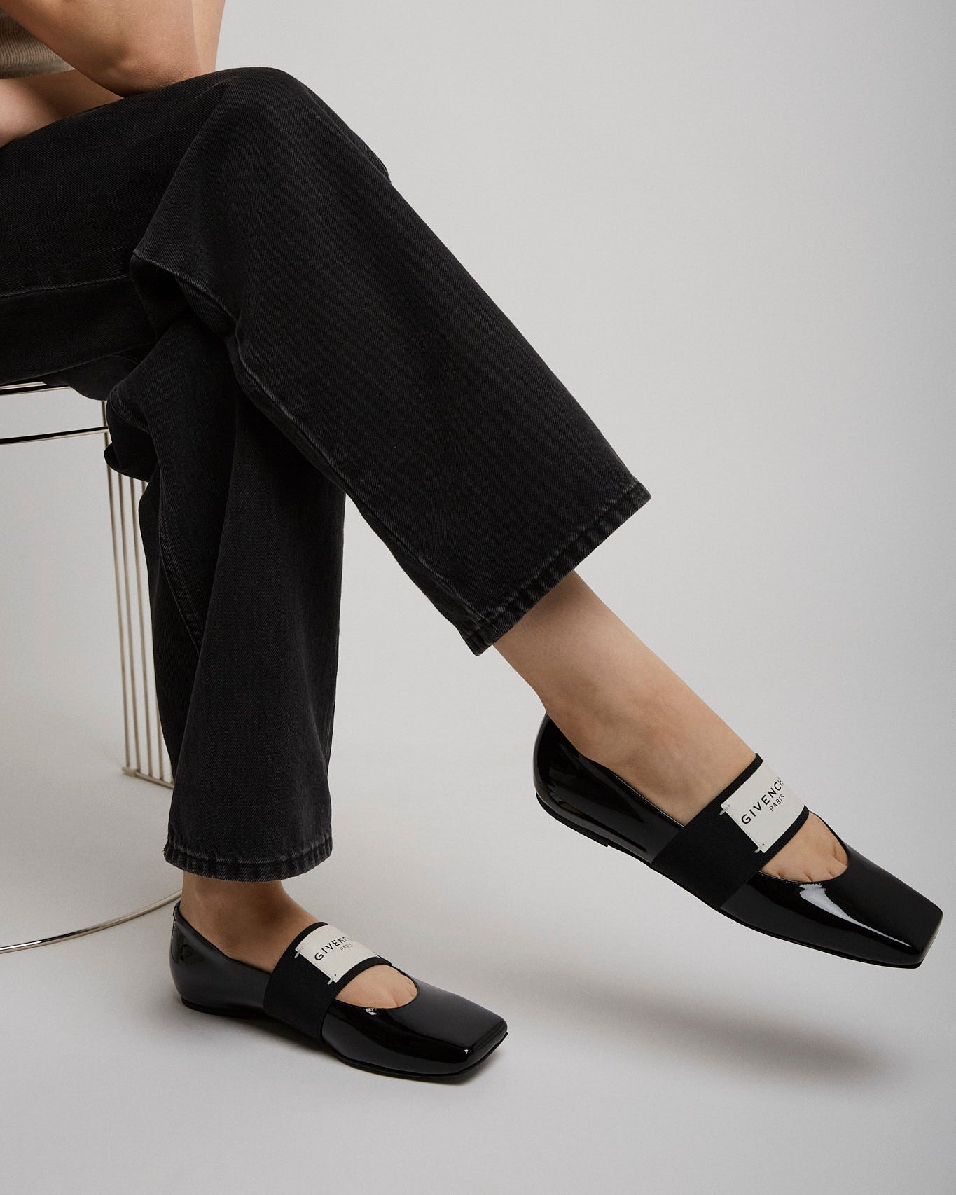 Person wearing black patent leather shoes with a white brand logo, sitting on a chair.