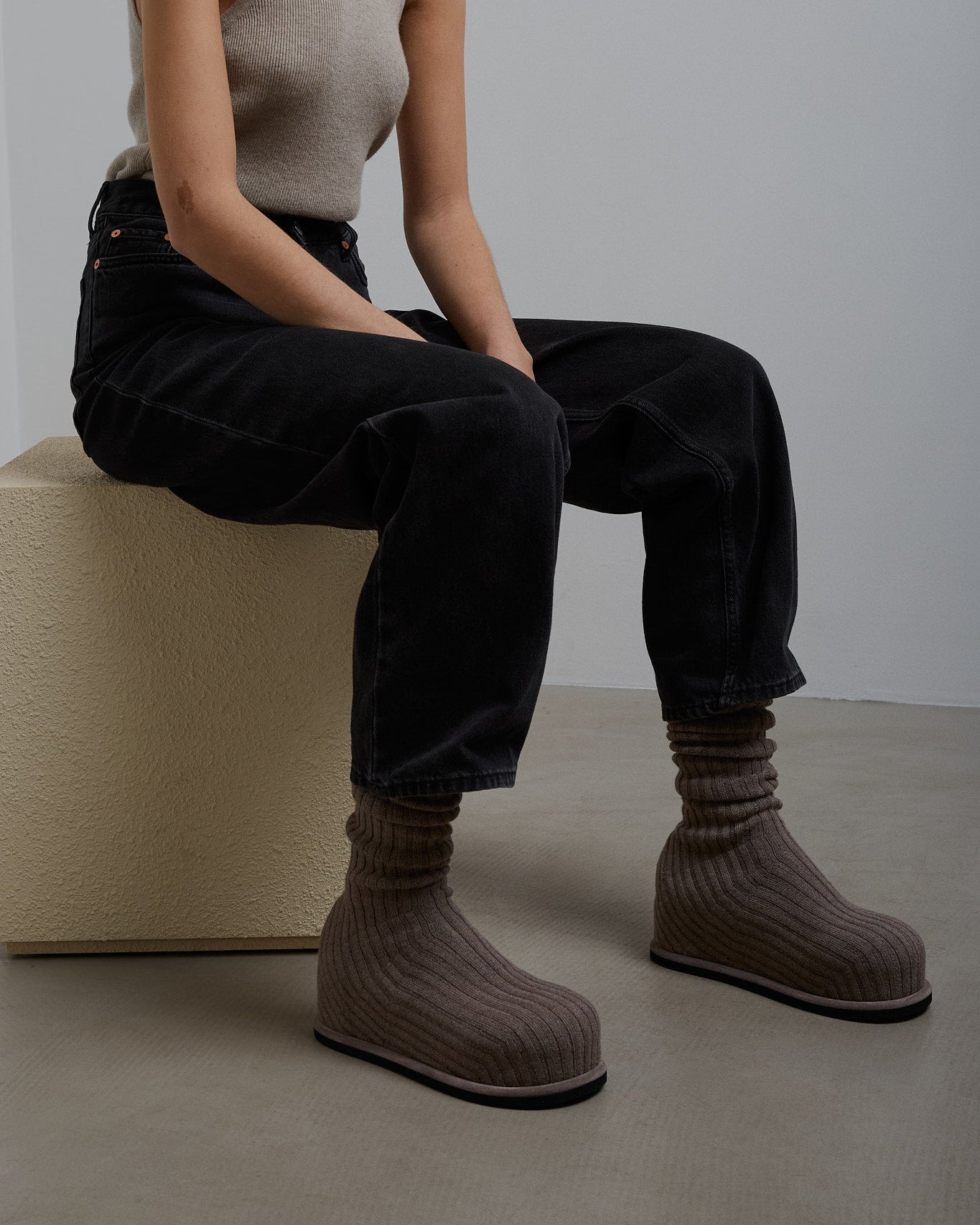 Person wearing brown knit boots sitting on a white block against a plain background