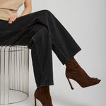 Person wearing a beige shirt, black jeans, and brown high-heeled shoes sitting on a wire stool.