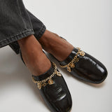 Black loafers with gold chain details worn with gray jeans on a light background