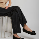 Person wearing black flats with a bow, sitting on a wire stool against a plain background