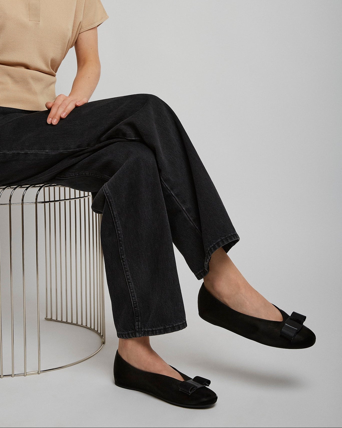 Person wearing black flats with a bow, sitting on a wire stool against a plain background