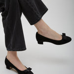 Person wearing black shoes with bows and black pants on a light gray background