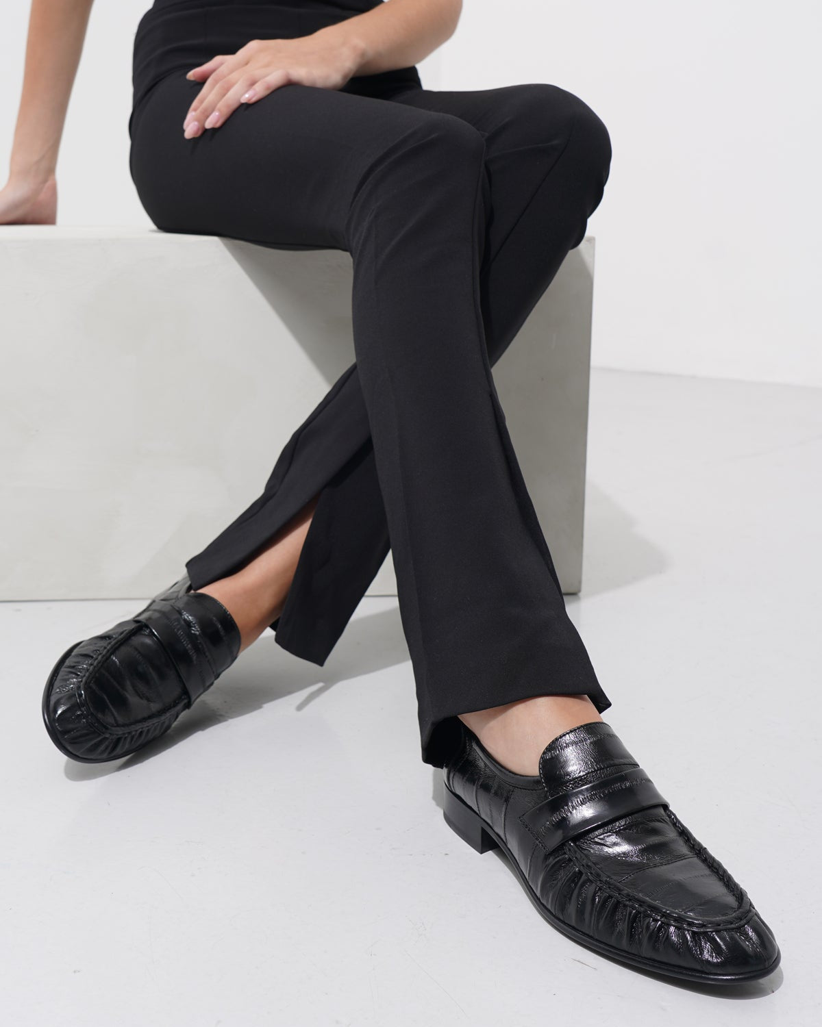 Person wearing black pants and shoes sitting on a white stool against a white background