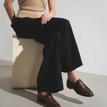 Person wearing a beige top and black wide-leg pants with brown shoes, sitting on a white box.