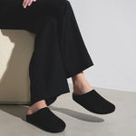 Person wearing black slippers and wide-leg pants sitting on a chair against a plain background