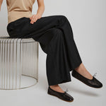 Person wearing a beige shirt, black pants, and black shoes sitting on a wire stool against a plain background.