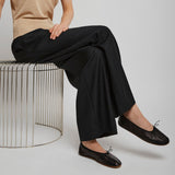 Person wearing a beige shirt, black pants, and black shoes sitting on a wire stool against a plain background.