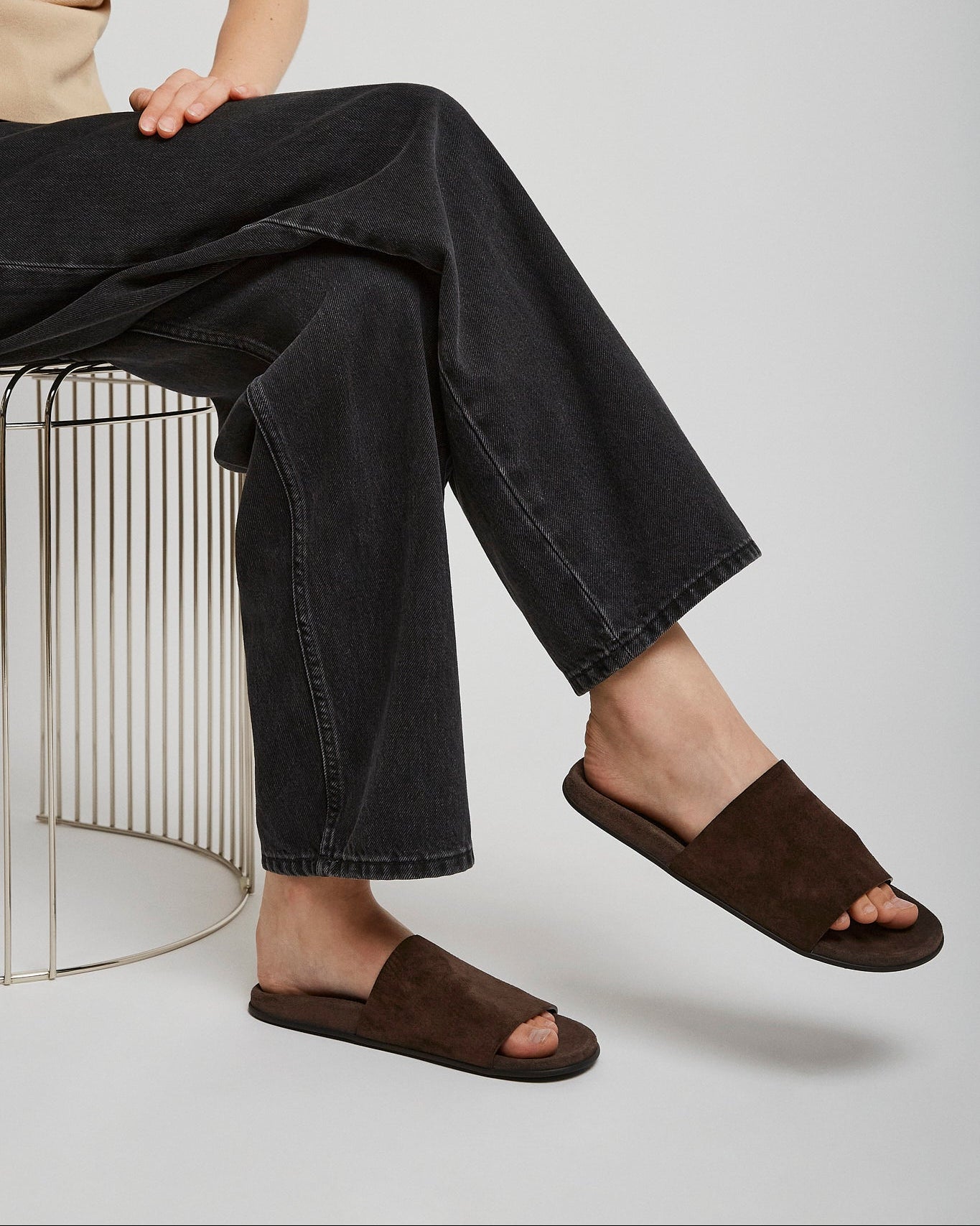 Person wearing brown sandals and black jeans sitting on a metal stool against a white background