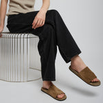 Person wearing a beige tank top, black pants, and brown sandals sitting on a white stool.