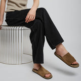 Person wearing a beige tank top, black pants, and brown sandals sitting on a white stool.