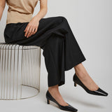 Person wearing a beige top and black pants sitting on a wire stool against a plain background