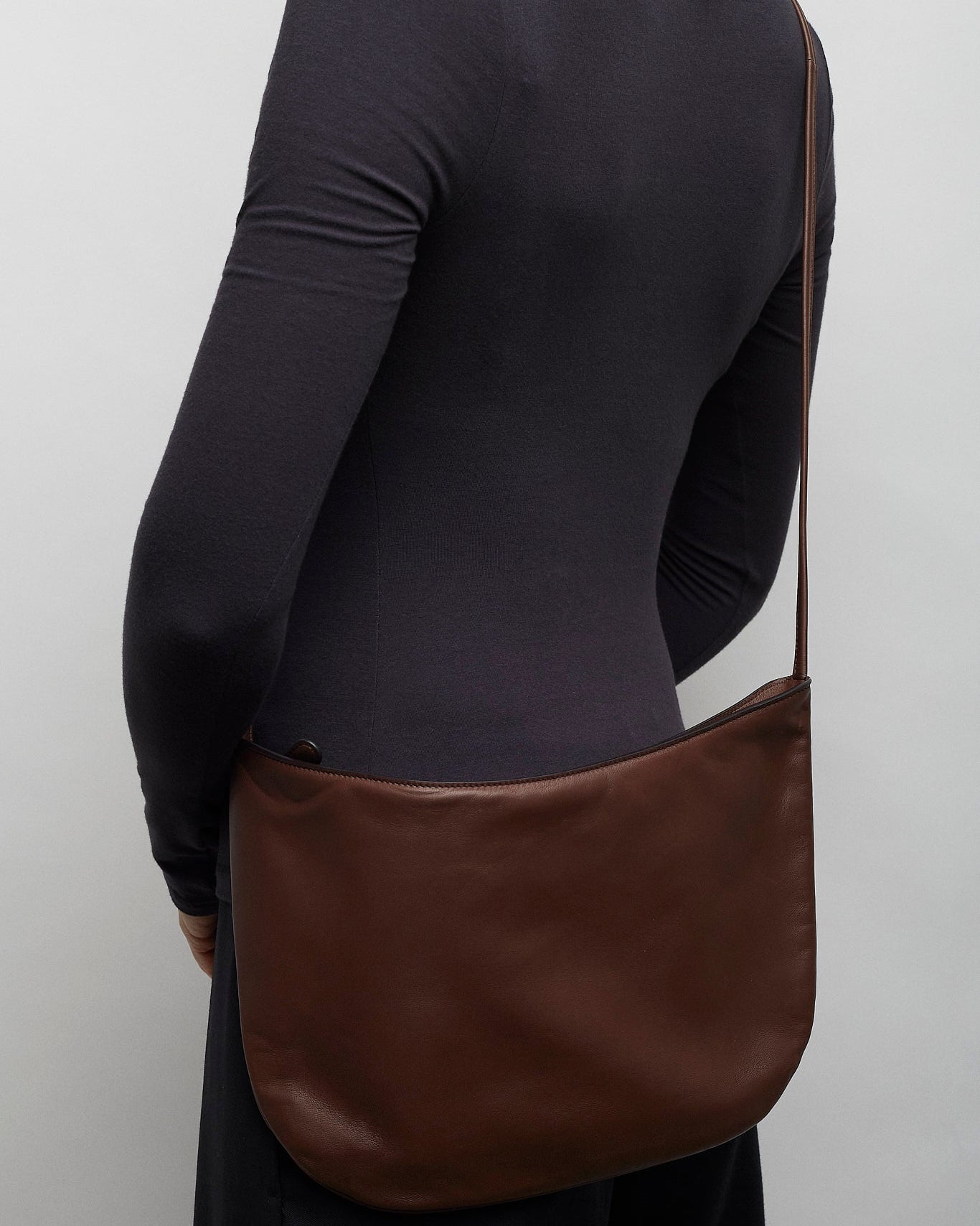 Person wearing a black long-sleeve shirt with a brown leather bag over one shoulder against a plain background.