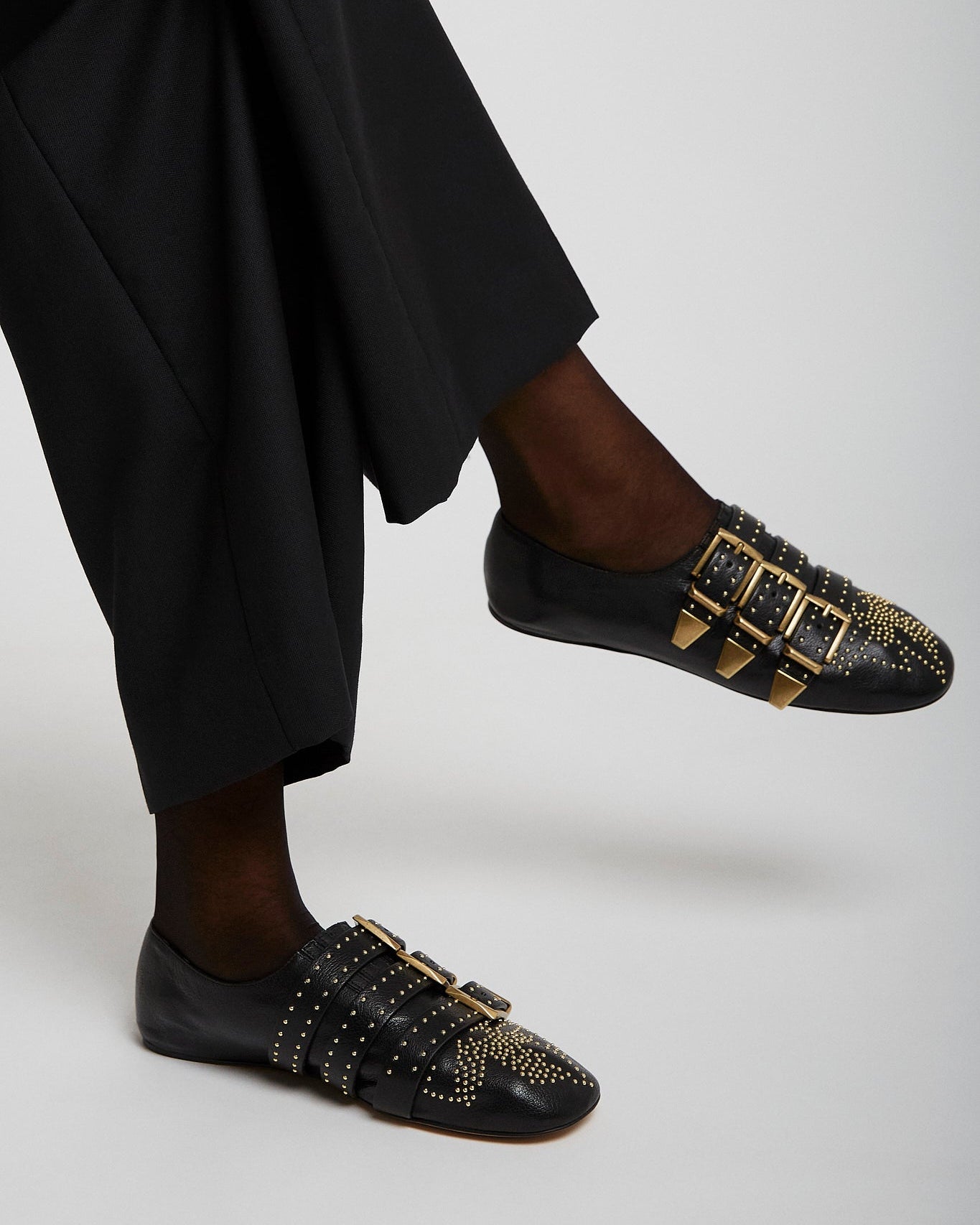 Black loafers with gold buckles worn by a person in black pants on a light gray background