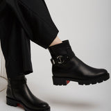 Black leather boots with a brand logo worn by a person sitting on a white chair.