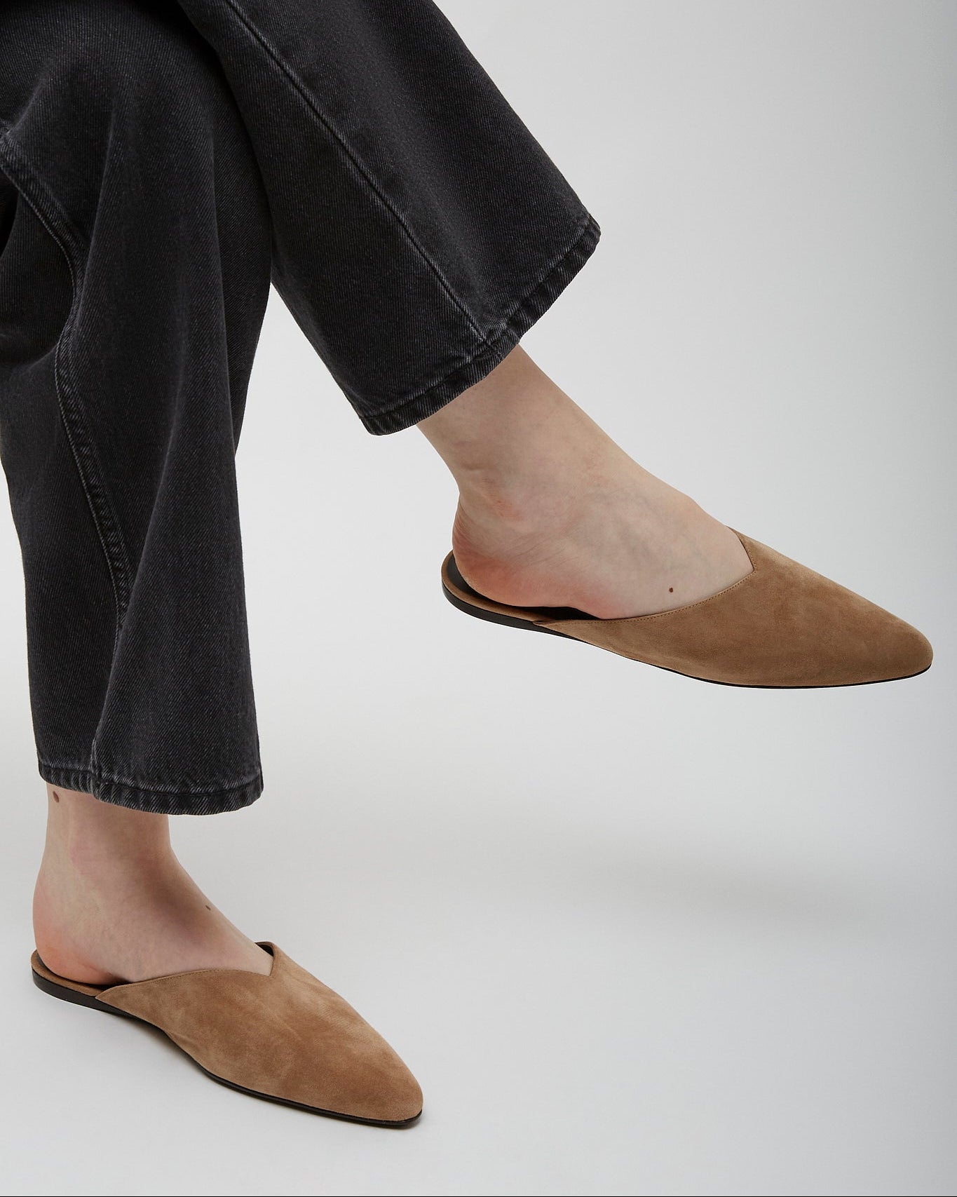 Person wearing brown suede slippers and black jeans on a light gray background