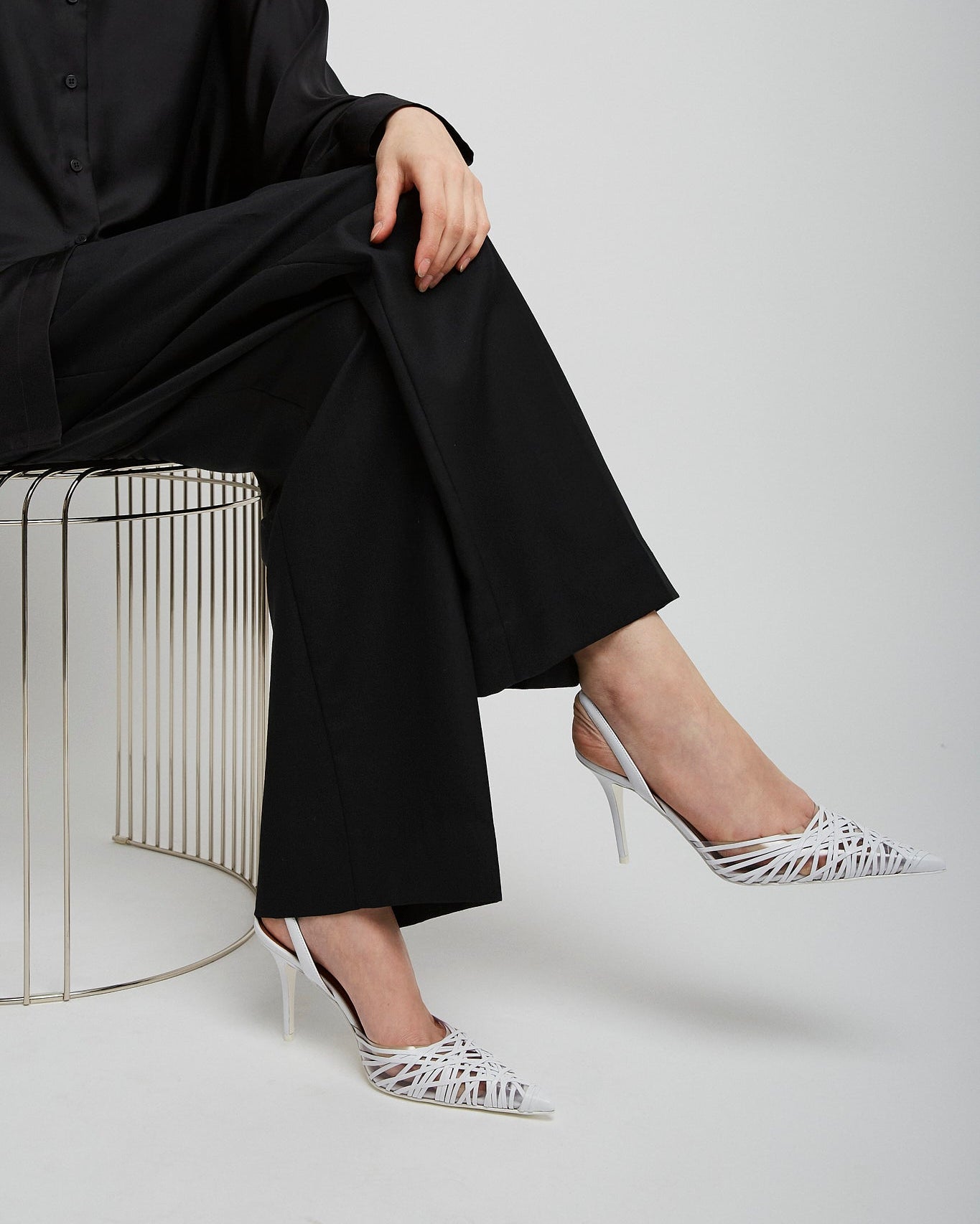 Person wearing a black outfit with white high-heeled shoes sitting on a metal stool against a white background